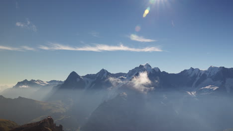 Una-Cautivadora-Toma-Aérea-Tomada-Por-Un-Dron-Muestra-Un-Panorama-Impresionante-De-Una-Cadena-Montañosa-Envuelta-En-Niebla,-Cielos-Azules-Y-El-Majestuoso-Sol-Brillando-Sobre-Ella.