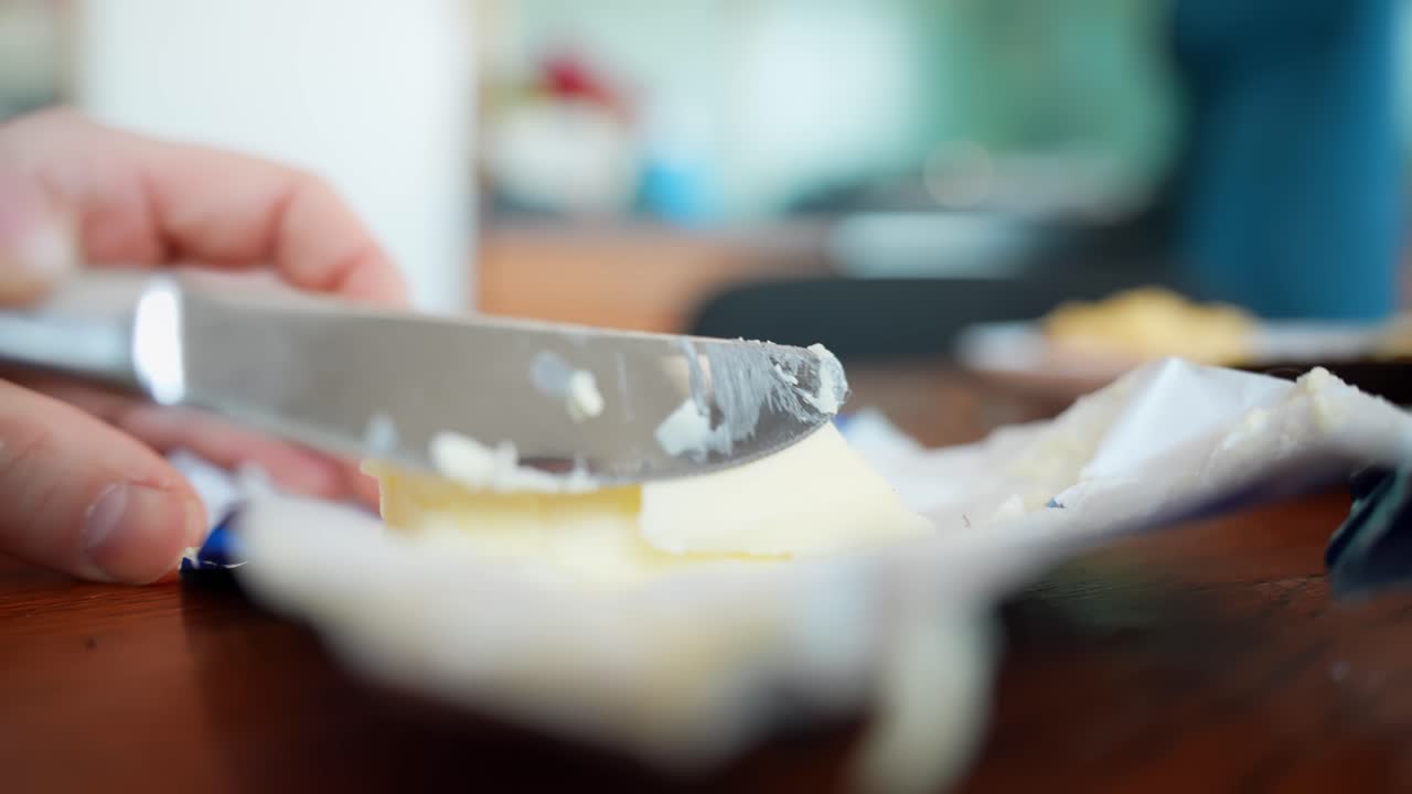 Close-up hand scoops butter from an opened wrapper with a knife on a wooden table, preparing ingredients for breakfast toast. Natural light food b-roll