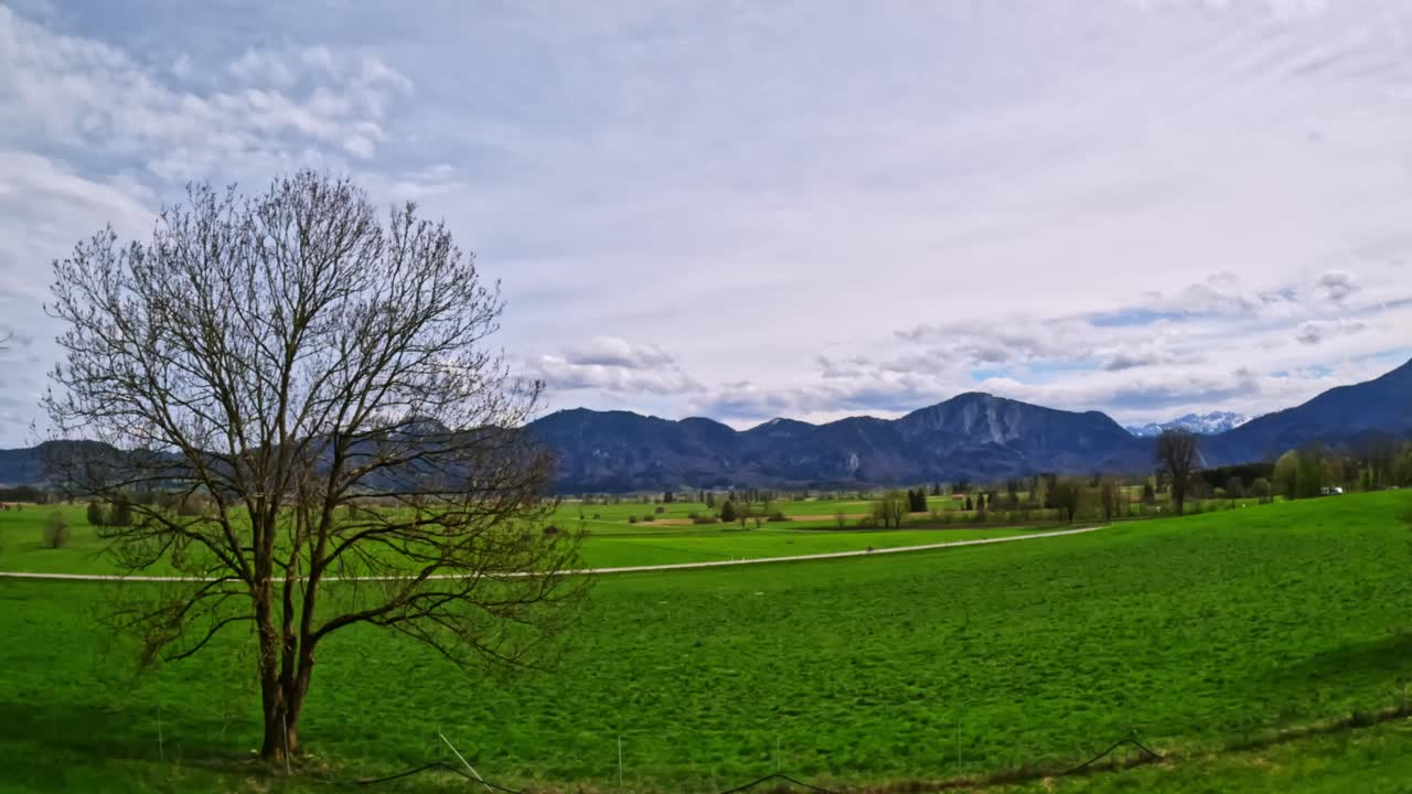 Wide open springtime meadow stretches toward distant Bavarian Alps with gently rolling hills and a cluster of scattered farmhouses, capturing the lush seasonal fields and tranquil rural landscape