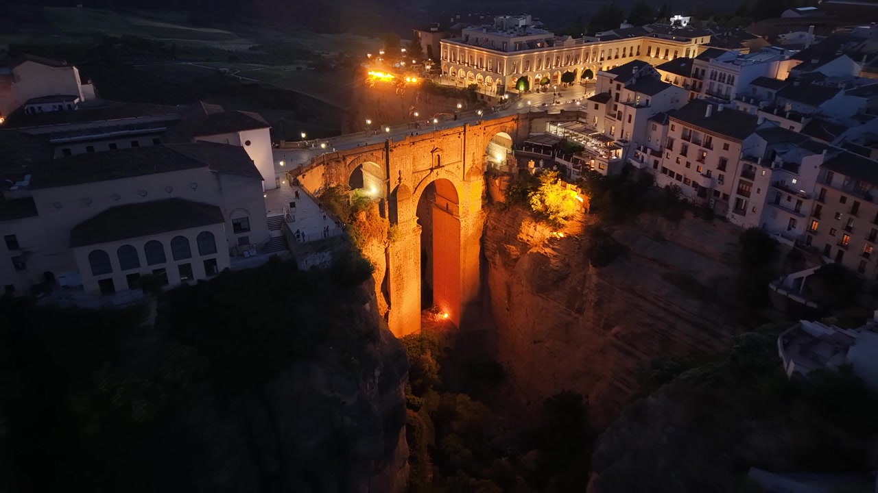 Vista aérea cinematográfica de dron del iluminado Puente Nuevo en Ronda, España, a la hora azul, con luces brillantes que resaltan el hito histórico sobre el desfiladero de El Tajo.