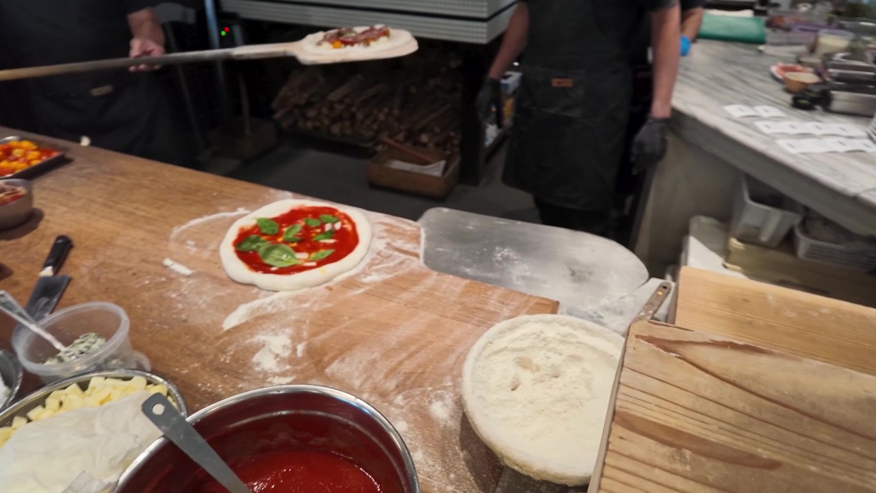 Ein Koch in schwarzer Schürze schiebt mit einem Holzschieber Pizza in den Ofen.