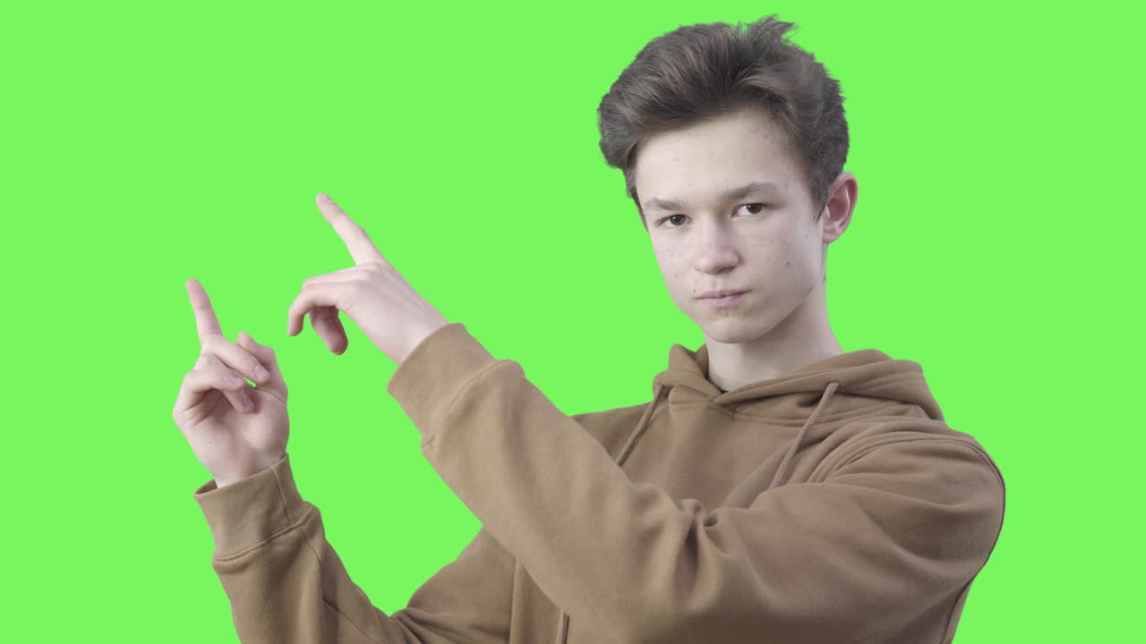 젊은 백인 소년은 손가락을 올리고 심각한 표정으로 카메라를 바라보고 있습니다. 십대 학생은 그린 스크린 배경에서 무언가를 보여주고 있습니다.