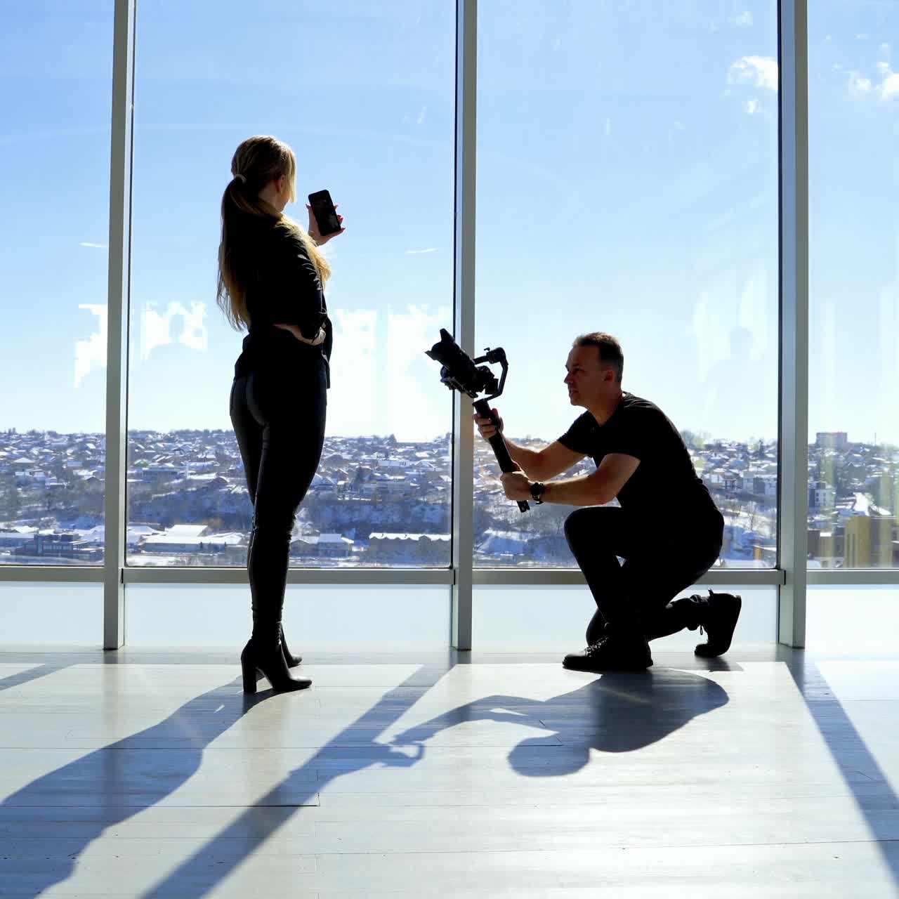 Photographer and a model with a phone indoors. Young woman doing selfie photos on her phone while man taking photos of woman on a professional camera