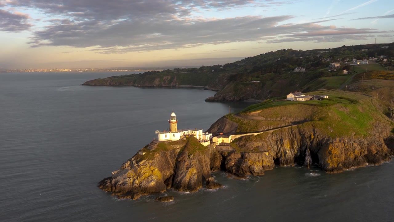 Il Faro di Baily e le case vicine si ergono su ripide scogliere costiere mentre un drone sorvola la baia nella calda luce della sera.