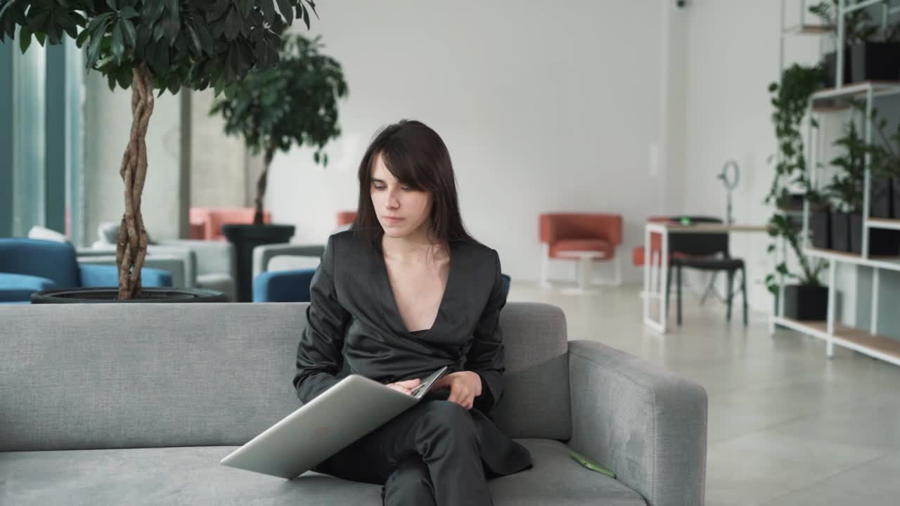 young tense woman, sitting on the couch in the business center's lobby with a laptop, bursts with anger and throws the laptop, contemplating problems