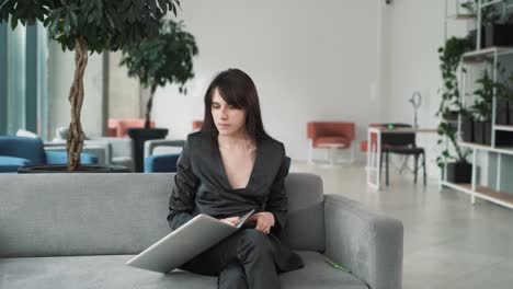 mujer joven tensa, sentada en el sofá en el vestíbulo del centro de negocios con una computadora portátil, estalla de ira y lanza la computadora portátil, contemplando problemas
