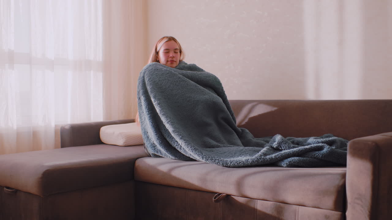 Woman sitting on sofa wrapping fluffy blanket around body with eyes closed in peaceful expression under warm morning light near window curtain, creating calm relaxed atmosphere inside cozy room