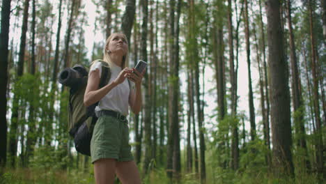 uma jovem mulher com um telemóvel caminha pela floresta viajando com uma mochila em câmera lenta. navegue pela floresta usando o navegador no seu telemóvel