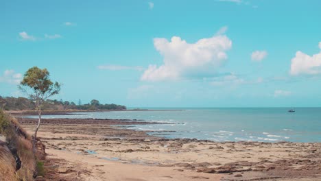 A-small-sized-sailing-boat-floats-over-beautiful,-tropical,-blue-water-just-off-the-coast-of-a-rocky-beach-made-of-rock,-stone,-sand-and-small-pebbles-under-a-beautifully-sunny-sky