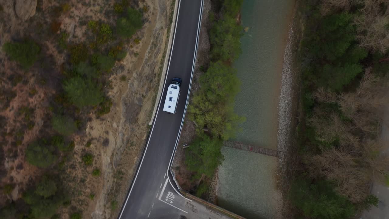 Top-down perspective of white integrated motorhome at a T-junction stop sign, crossing a narrow concrete bridge over a braided riverbed, navigating technical mountain passes
