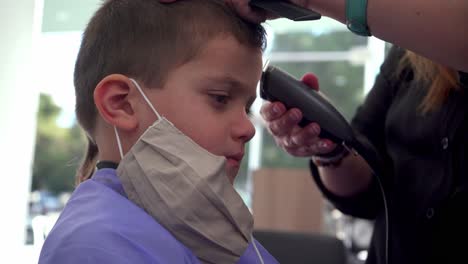 Cute-caucasian-boy-gets-a-haircut-at-hair-salon,-wearing-covid19-face-mask,-tight-static-shot