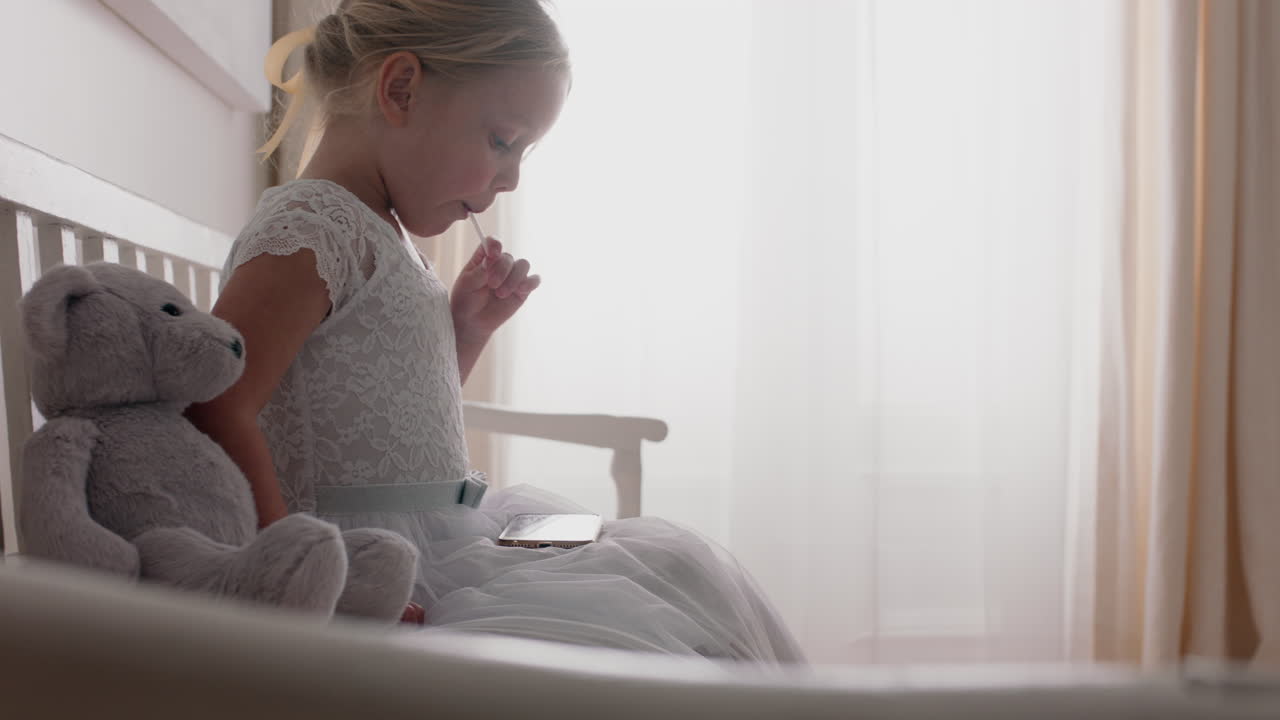 bellissima ragazzina che usa lo smartphone guardando intrattenimento online seduta su una panchina con un orsacchiotto che mangia un lecca-lecca a casa 4k