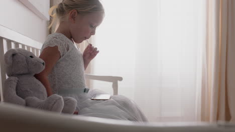 beautiful-little-girl-using-smartphone-watching-online-entertainment-sitting-on-bench-with-teddy-bear-eating-lollipop-at-home-4k-footage