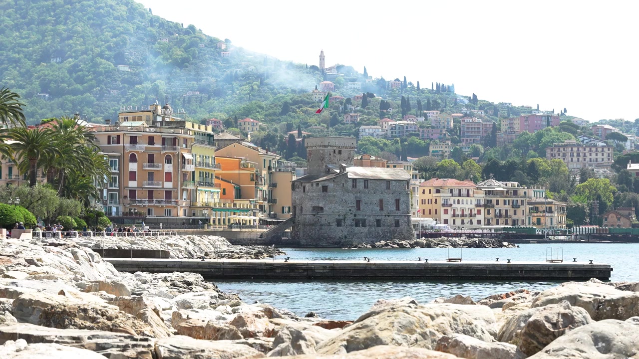 Vista grandangolare del Castello di Rapallo e della vivace architettura ligure in riva al mare sotto la luce del giorno intensa, con costa rocciosa, lussureggianti colline verdi e acqua blu limpida. Nessuna persona visibile