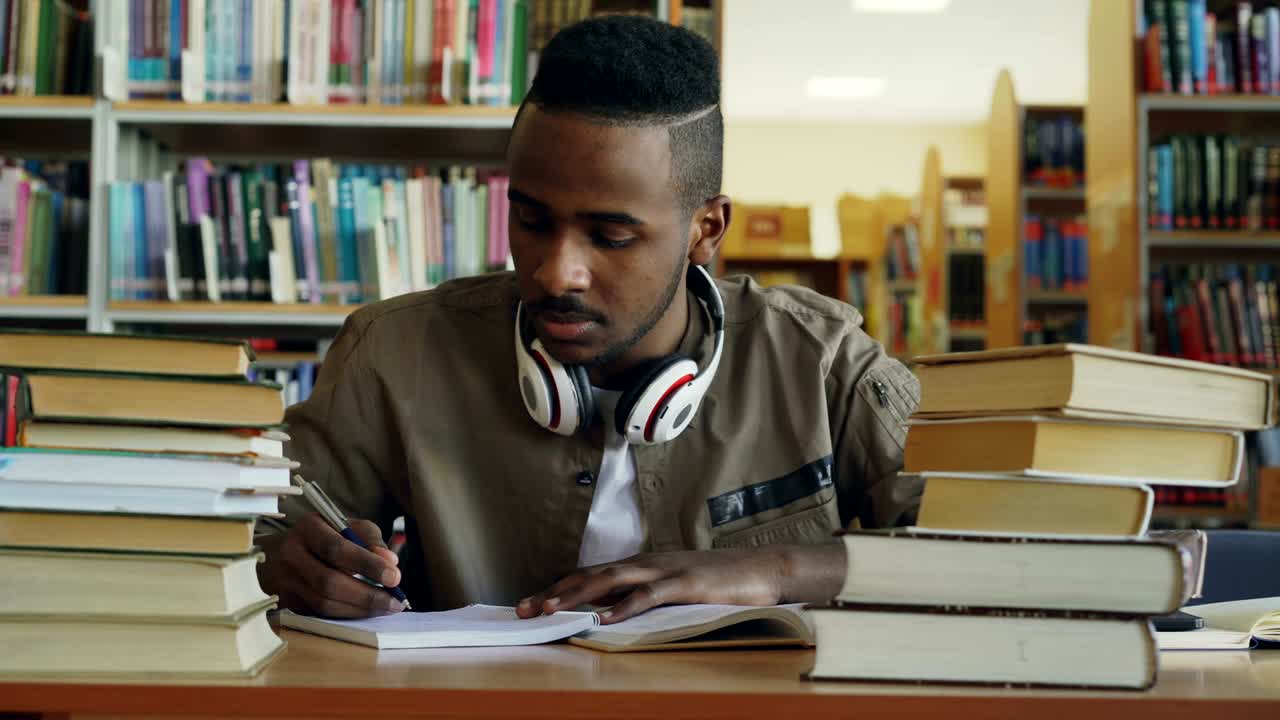 Positive african american young handsome guy with big headphones is sitting at table with books, looking through window, smiling and writing in his copybook