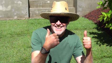 landscaper squatting with sunglasses, and a straw hat puts two thumbs up in completion in slow motion