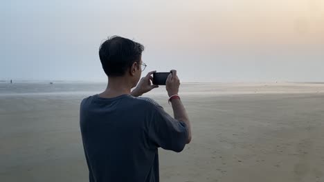 Vista-Lateral-En-Cámara-Lenta-De-Un-Hombre-Indio-Con-Camiseta-Azul-Parado-En-Una-Playa-Agradable-Haciendo-Clic-En-Fotos-De-Puesta-De-Sol