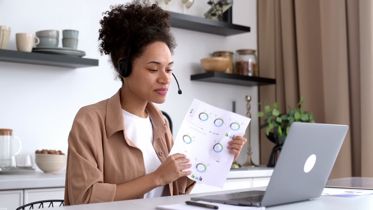 Friendly pretty, african american girl with headset, freelancer, financial advisor, sits at desk in the kitchen, talks via video communication with partners, discuss and showing financial charts,smile