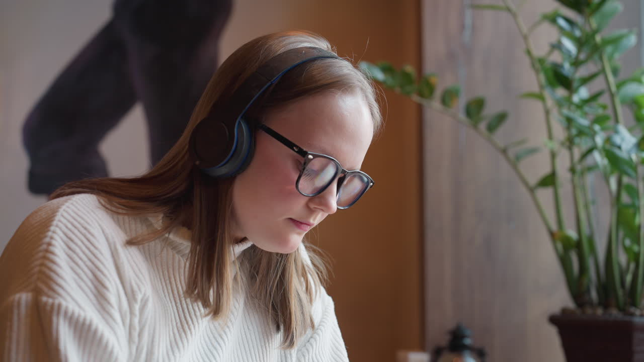 young woman wearing headset focuses deeply while sitting by window next to large potted plant, warm wall mural and cozy light create relaxed atmosphere as she enjoys audio in quiet space