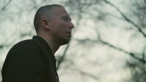 a man in a green jacket smoking a cigarette outdoors at dusk, the background is blurred with a soft, warm light