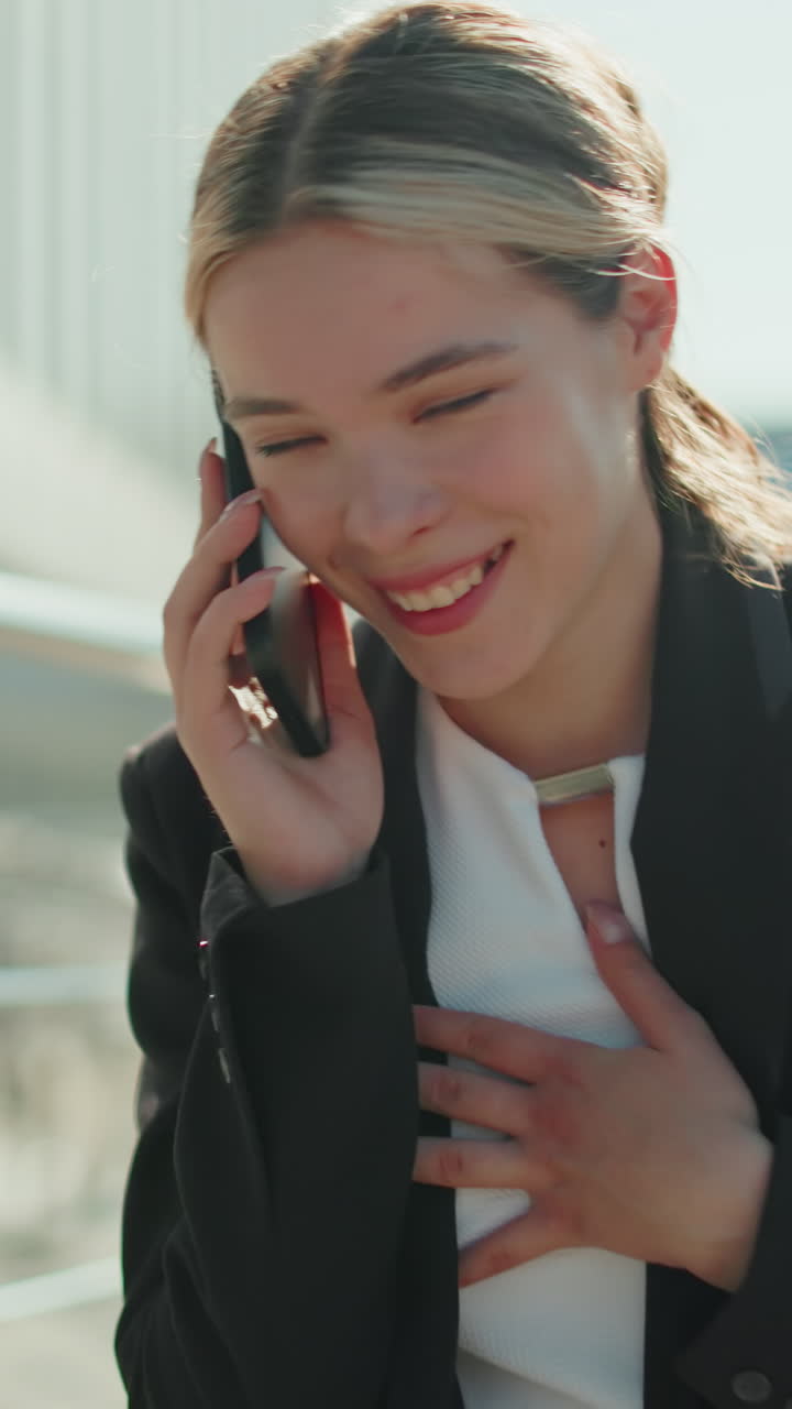 Cheerful lecturer in professional outfit walking confidently while talking on phone with friend, smiling with joy as person approaches from behind in urban setting with glass building background