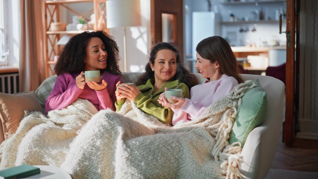 Diverse women comfortable rest on couch living room. Smiling friends drinking