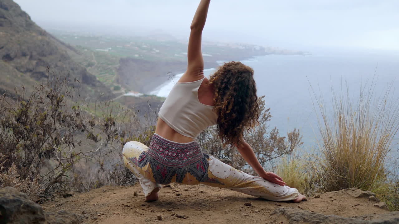 Perched on a cliff's edge, a woman assumes a warrior pose, raising her arms and breathing in the ocean air as she practices yoga