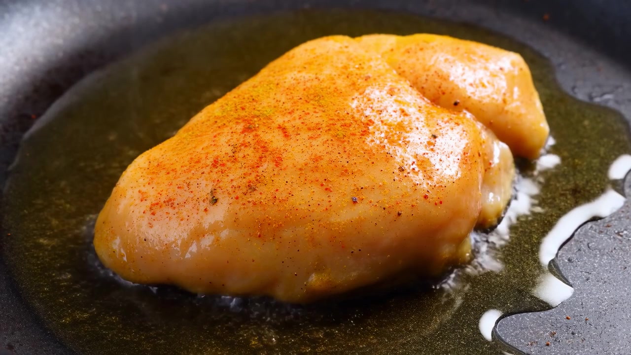 Pollo crudo disossato con panatura di spezie rosse e nere riposa in poco olio d'oliva all'interno di una padella antiaderente scura, creando riflessi lucidi, una consistenza ricca e una semplice disposizione degli ingredienti.