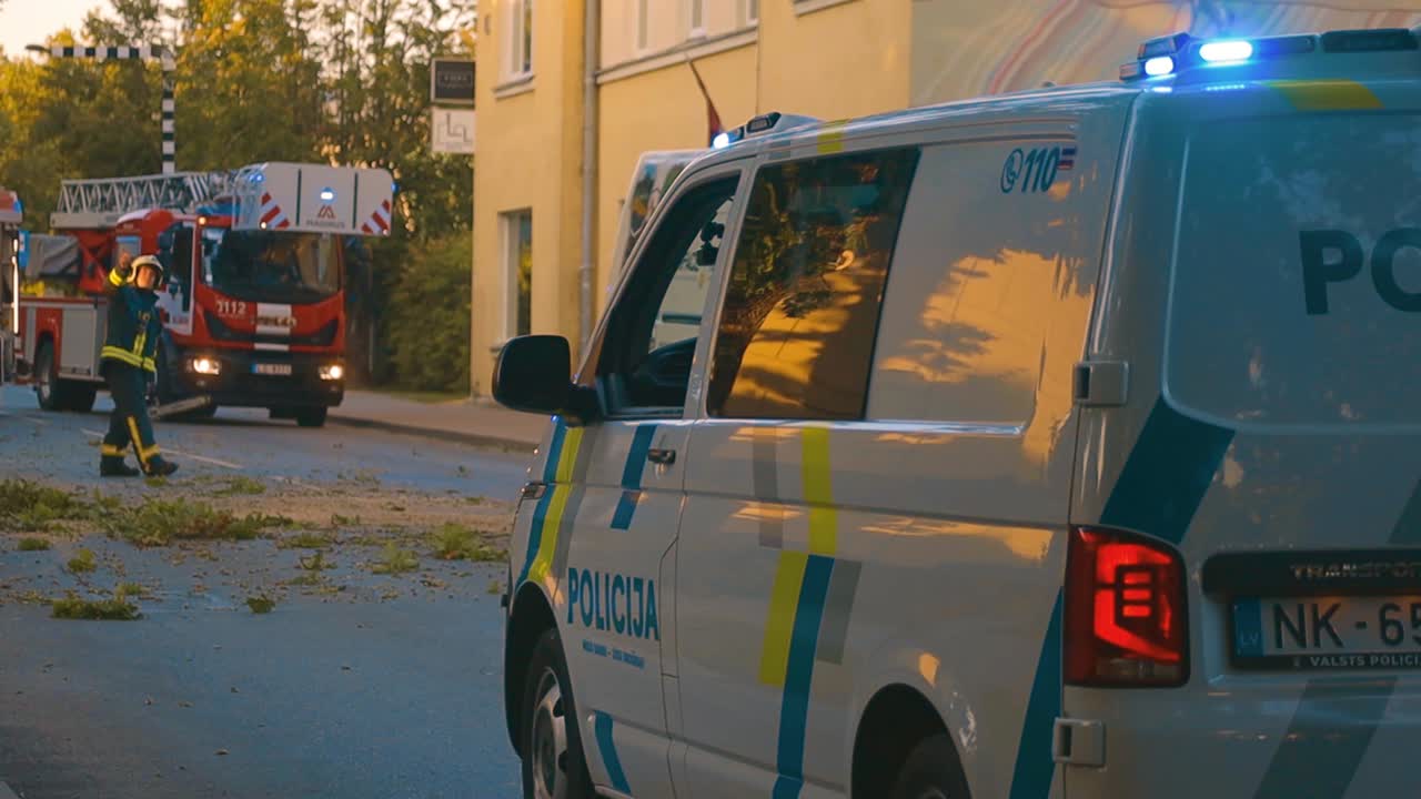 A police car with activated beacon lights is parked on a town road at night. Surrounding buildings and streetlights are visible, creating a calm urban atmosphere. Copy space available.