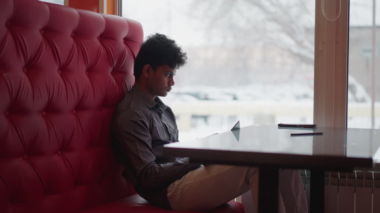 Student sitting alone on red cushioned bench with legs bent, using tablet at table beside large window during snowy day, focused on screen, studying or working in quiet cozy indoor environment