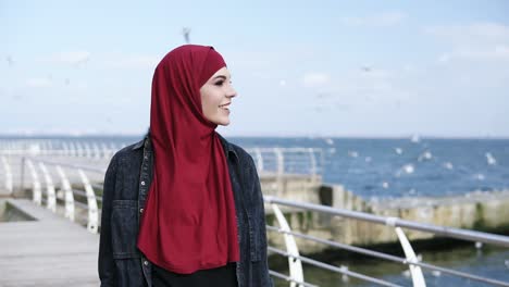 Young-attractive-muslim-girl-enjoys-having-a-walk-near-the-sea-side-with-seagulls-flying-around-on-the-background.-Outdoors-footage