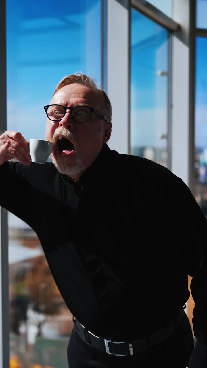 Old bearded grimacing man wearing black clothes. Smiling male pretends to bite a little coffee cup. Vertical video.