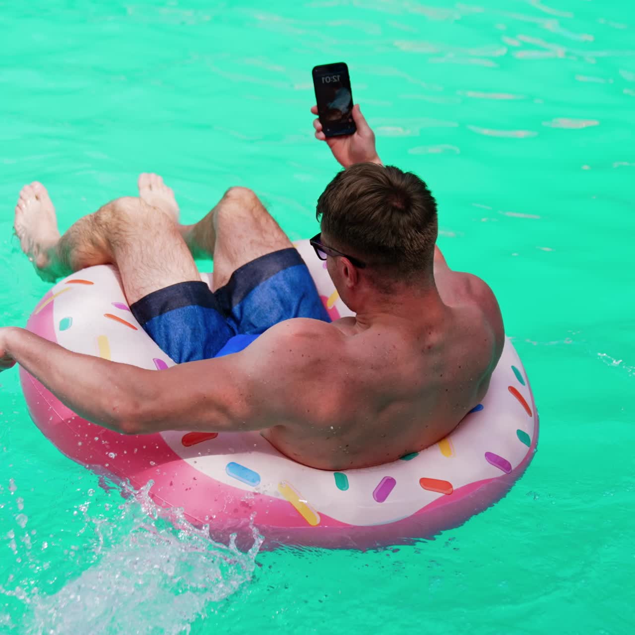 Muscular male sits in the inflatable circle holding a phone. Man taking selfie while resting in the pool