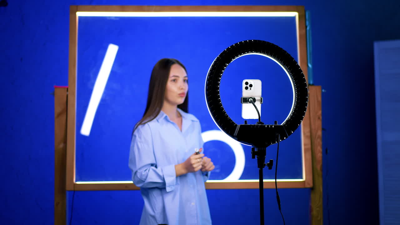 Young woman with ring light. A young woman stands confidently in front of a blue background, presenting while holding a smartphone