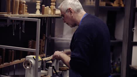 a senior man in eyeglasses standing in front equipment and cutting wooden knob out of wood piece spinning on machine using chisel, close up shot. slow motion. side view