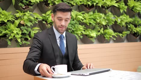 hombre de negocios guapo en traje formal escribiendo en la pantalla de la computadora portátil y bebiendo una taza de café en un restaurante.