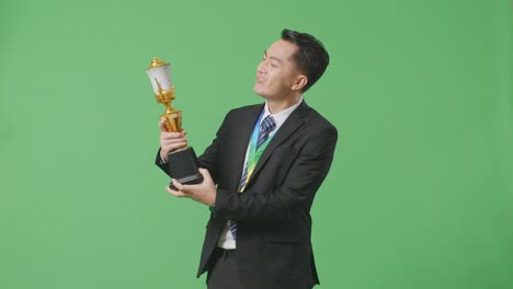 hombre de negocios asiático en traje y corbata con una medalla de oro besando un trofeo de oro estando orgulloso de ganar como el primer ganador en el fondo de la pantalla verde en el estudio