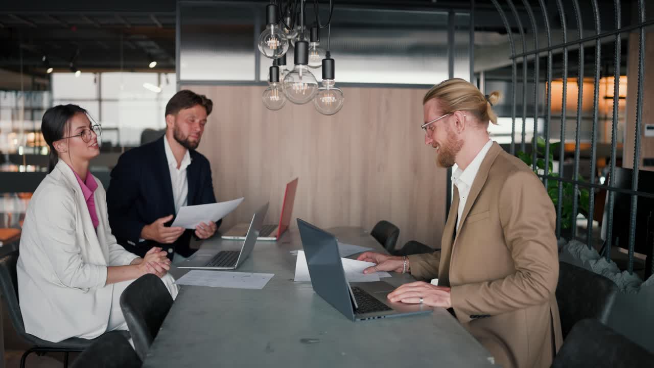 A brunette girl in a white suit gives a blonde man in a light brown jacket papers for work. Paper work in modern office. A blond man with a beard and glasses communicates with his work colleagues at a table in the office