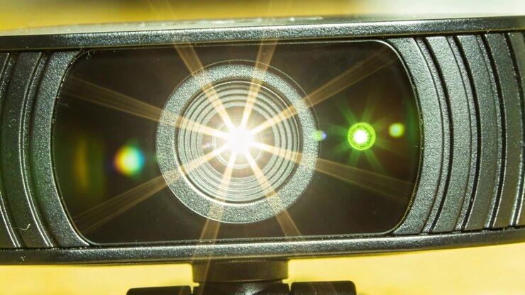 Viser USB webcam-linse, der reflekterer lys uden for billedet ved skrivebord, producerer stjerneblænding og grøn LED.