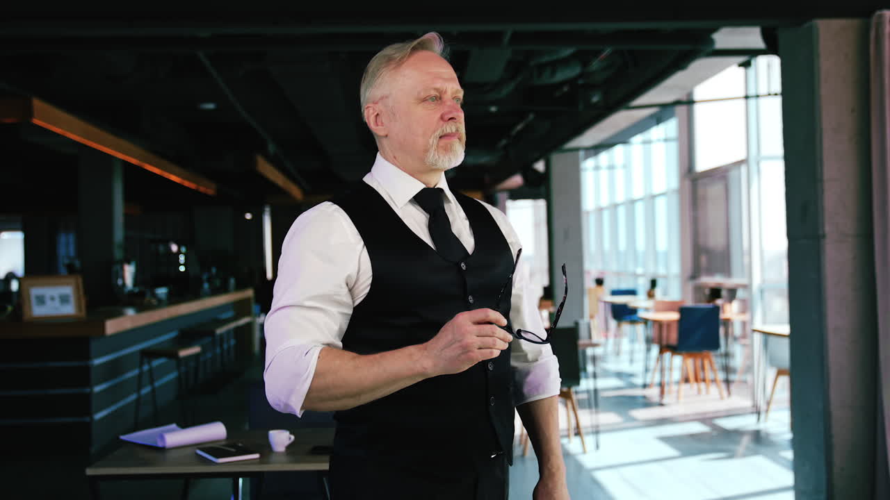 Bearded Caucasian old man in white shirt and black vest standing indoors. Portrait of businessman holding glasses and looking aside and then to the camera.
