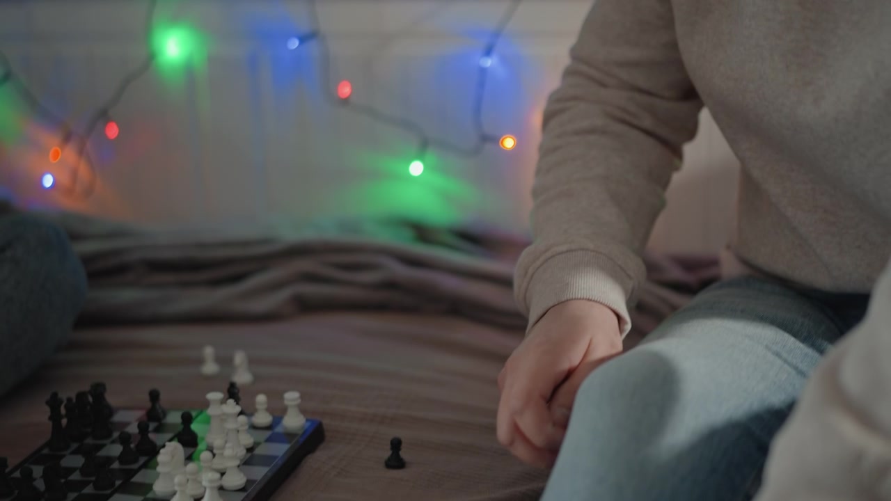 Due persone su una coperta giocano a scacchi in modo informale al chiuso, mentre uno si sporge in avanti per raggiungere un pezzo, creando un momento di concentrazione, interazione, luce soffusa e un rilassato svago domestico, con scacchi, gioco e scacchiera.