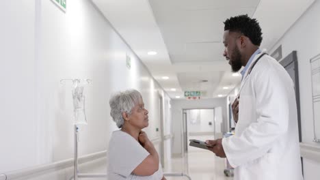serious diverse male doctor with tablet talking to senior patient on bed in slow motion, unaltered