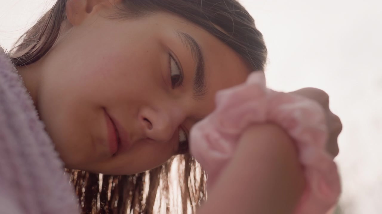 Young woman with long brown hair gently dries her wet strands in soft natural light wearing purple towel. Her focused expression and texture of damp locks are highlighted by warm sunlight