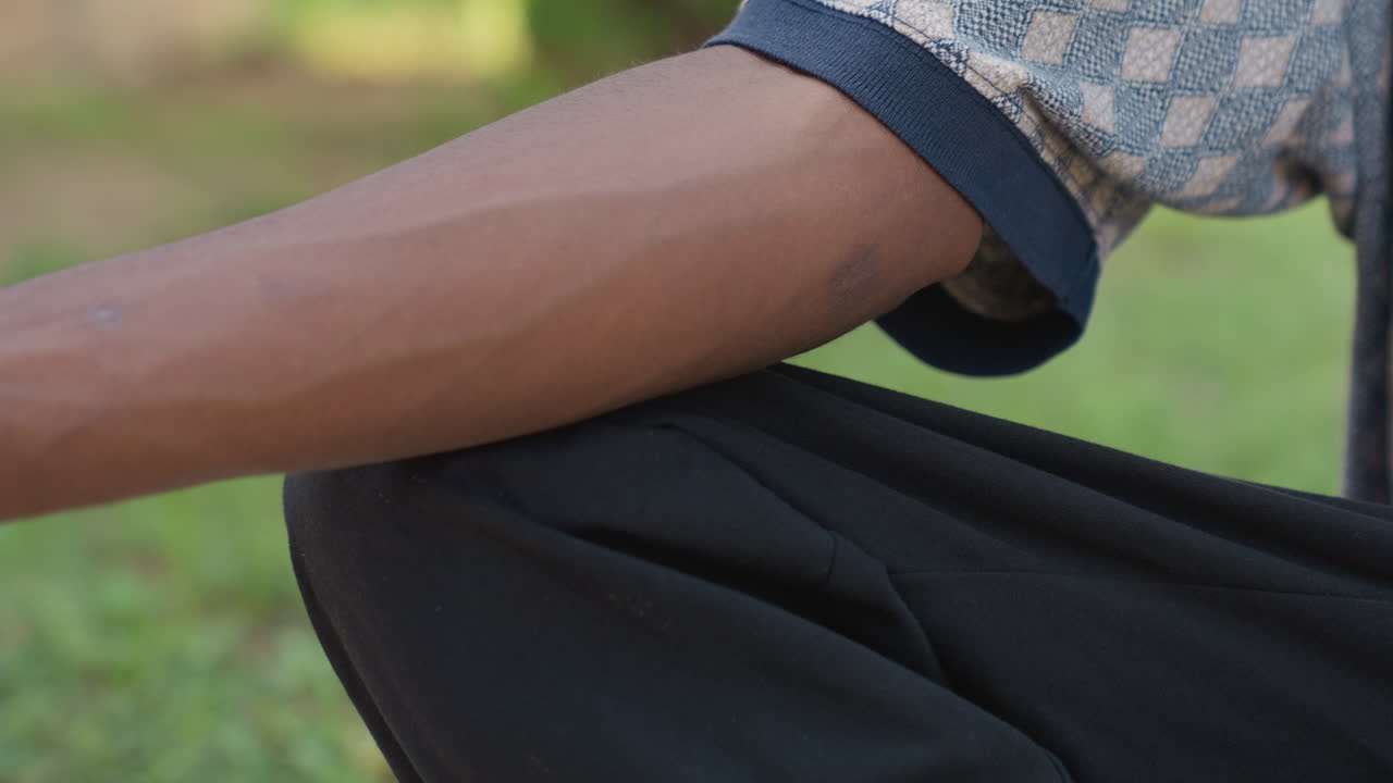 Schwarzer Athlet dehnt den Arm im Freien, sitzt im Schneidersitz auf Gras im Park, trägt schwarze Hose und Kurzarmshirt. Unterarmvenen und Muskeln sind betont. Eine ruhige Aufwärmroutine vor dem Training, natürlich.