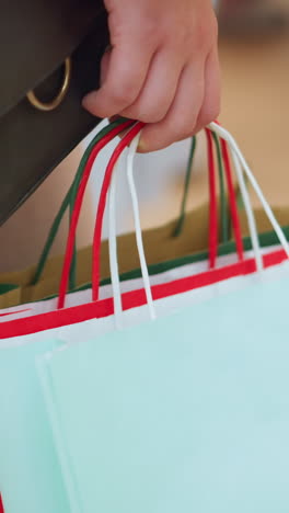 vista de la pierna de una dama sosteniendo tres bolsas de compras coloridas en rojo, verde y verde menta, con un bolso de mano negro, de pie quieto, fondo borroso incluye otras personas en un entorno de compras