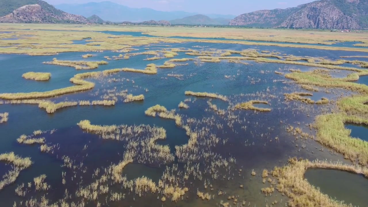 Panoraamainen korkea ilmakuva Dalyan-joen suiston kultaisten ruovikkojen ja sinisten vesikanavien labyrintista, jota kehystävät vuoret Muglassa, Turkissa.