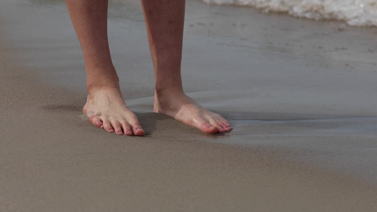 Close up of slim tanned legs of a young woman standing on the seashore. Waves wash her feet
