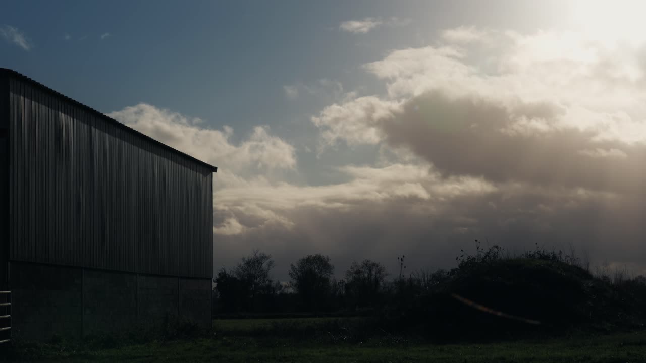 Un'inquadratura ampia di un fienile agricolo in lamiera ondulata grigia che si erge in un campo verde sotto un cielo cupo e nuvoloso. Paesaggio rurale cinematografico in campagna