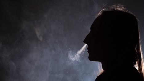 vista lateral de primer plano de silueta aislada de un hombre con el pelo largo fumando en una habitación oscura que sopla humo por la nariz