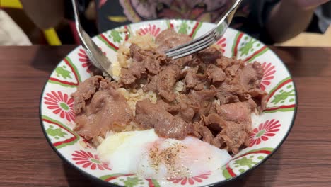 a person uses a spoon and fork to mix sliced beef, steamed rice, and a soft-cooked egg in a patterned bowl under bright indoor lighting